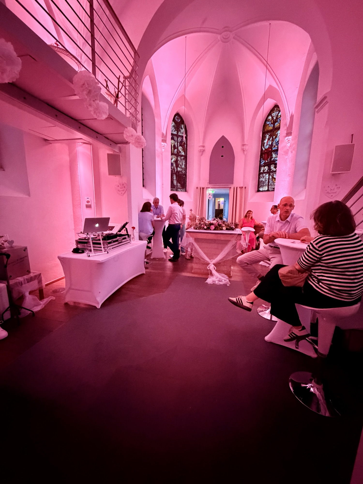 Innenraum einer historischen Kirche mit rosa Eventbeleuchtung und Spitzbogenfenstern, Stehtische mit Hochzeitsgästen und DJ-Tisch am Rand