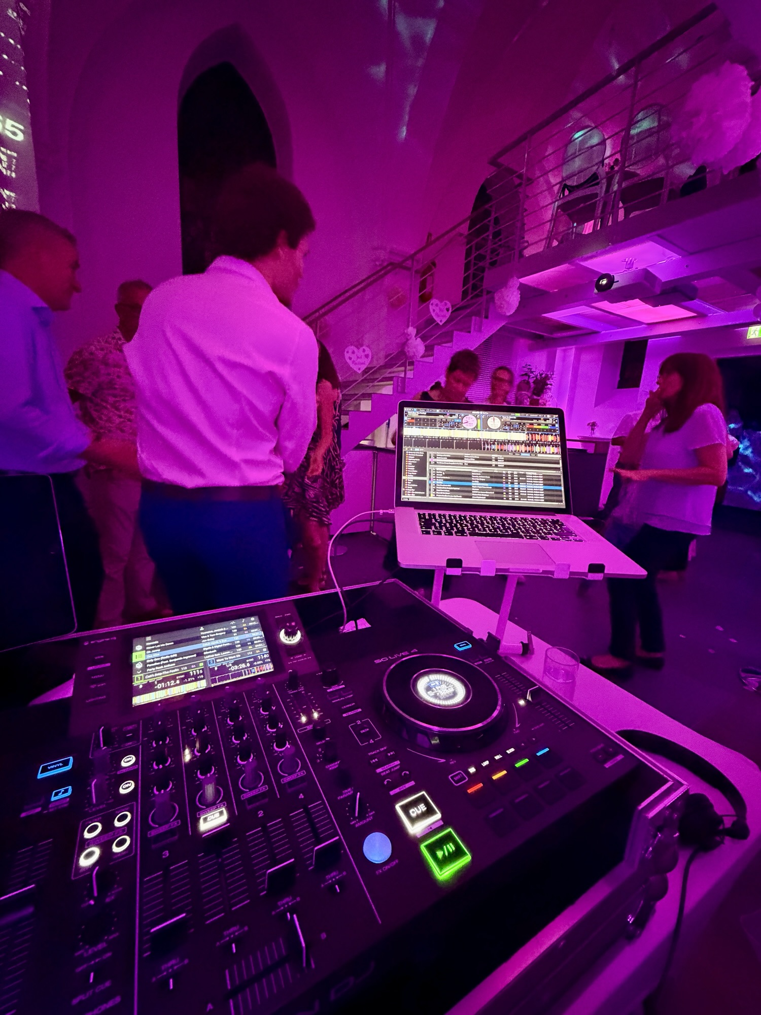 DJ-Equipment mit Laptop und Denon-Controller in einer Bar mit violetter Eventbeleuchtung, im Hintergrund tanzende Gäste