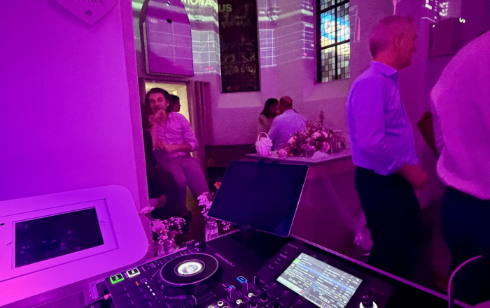 DJ-Pult in einer Kirche mit gotischem Gewölbe, bunte Videoprojektion an der Wand, im Hintergrund Hochzeitsgäste und ein 'Just Married'-Schild