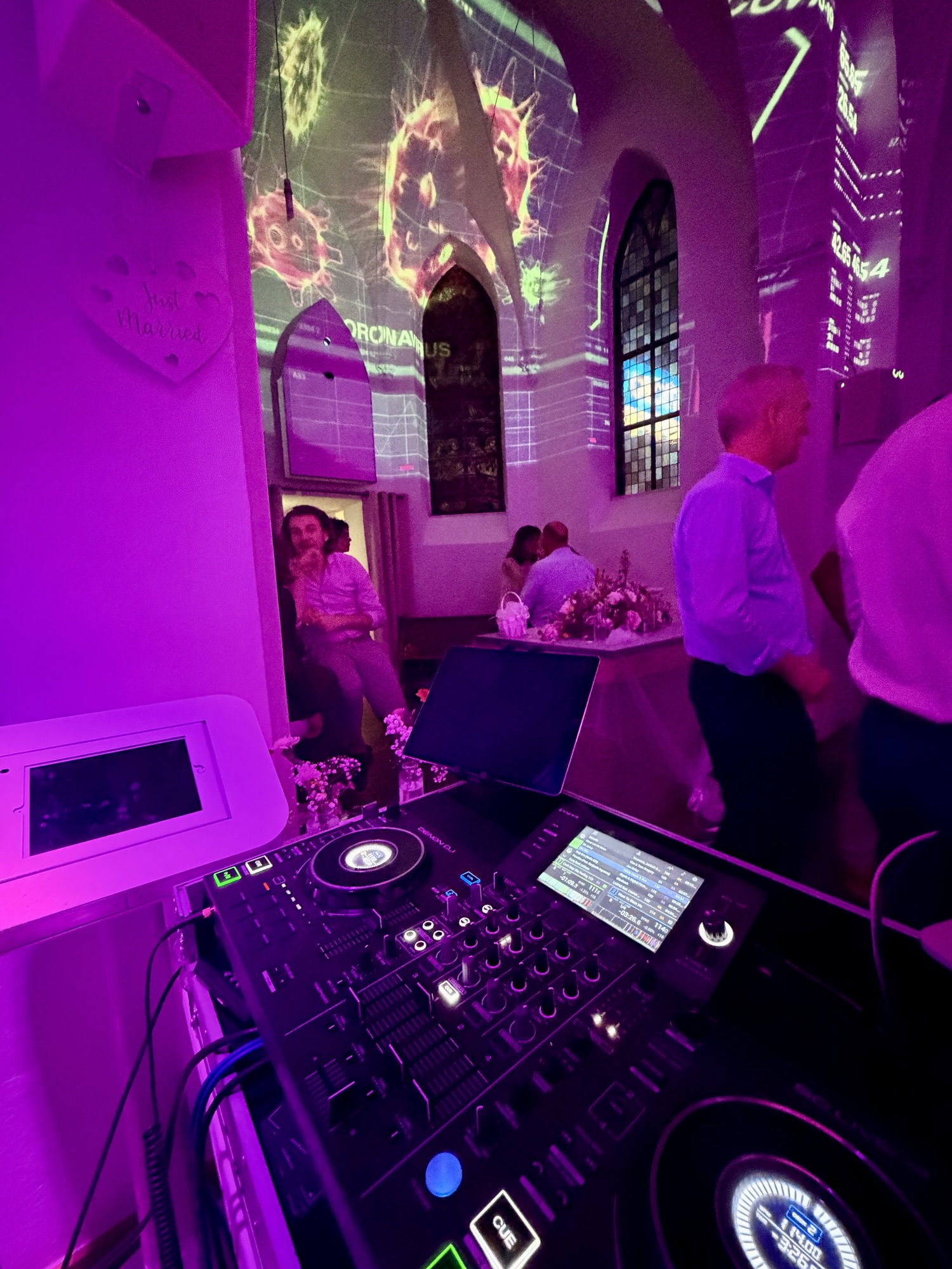DJ-Pult in einer Kirche mit gotischem Gewölbe, bunte Videoprojektion an der Wand, im Hintergrund Hochzeitsgäste und ein 'Just Married'-Schild