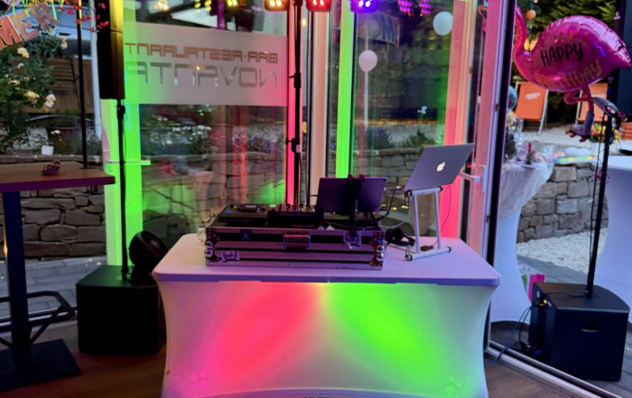 Aufgebautes DJ-Pult mit roter, grüner und lila LED-Beleuchtung unter einem Partyzelt, daneben ein pinker Flamingo-Luftballon mit Happy-Birthday-Schriftzug