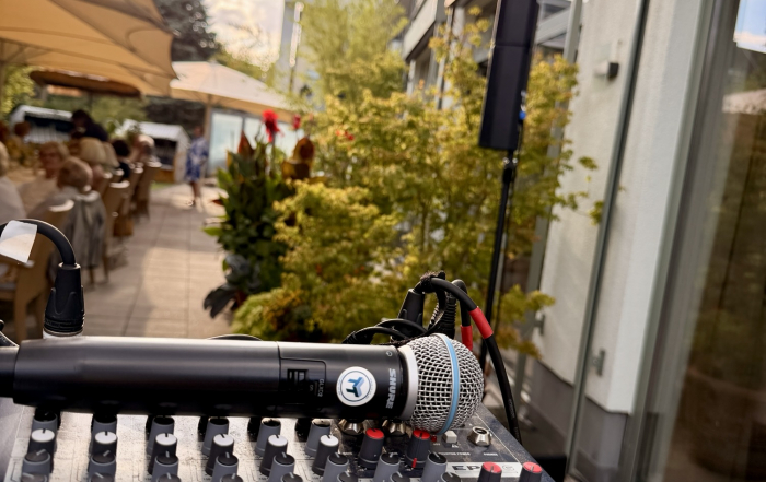 Blick über ein Mischpult mit Shure-Handmikrofon auf eine Restaurant-Terrasse mit Sonnensegeln bei Abendlicht, im Hintergrund die Fassade des Gebäudes