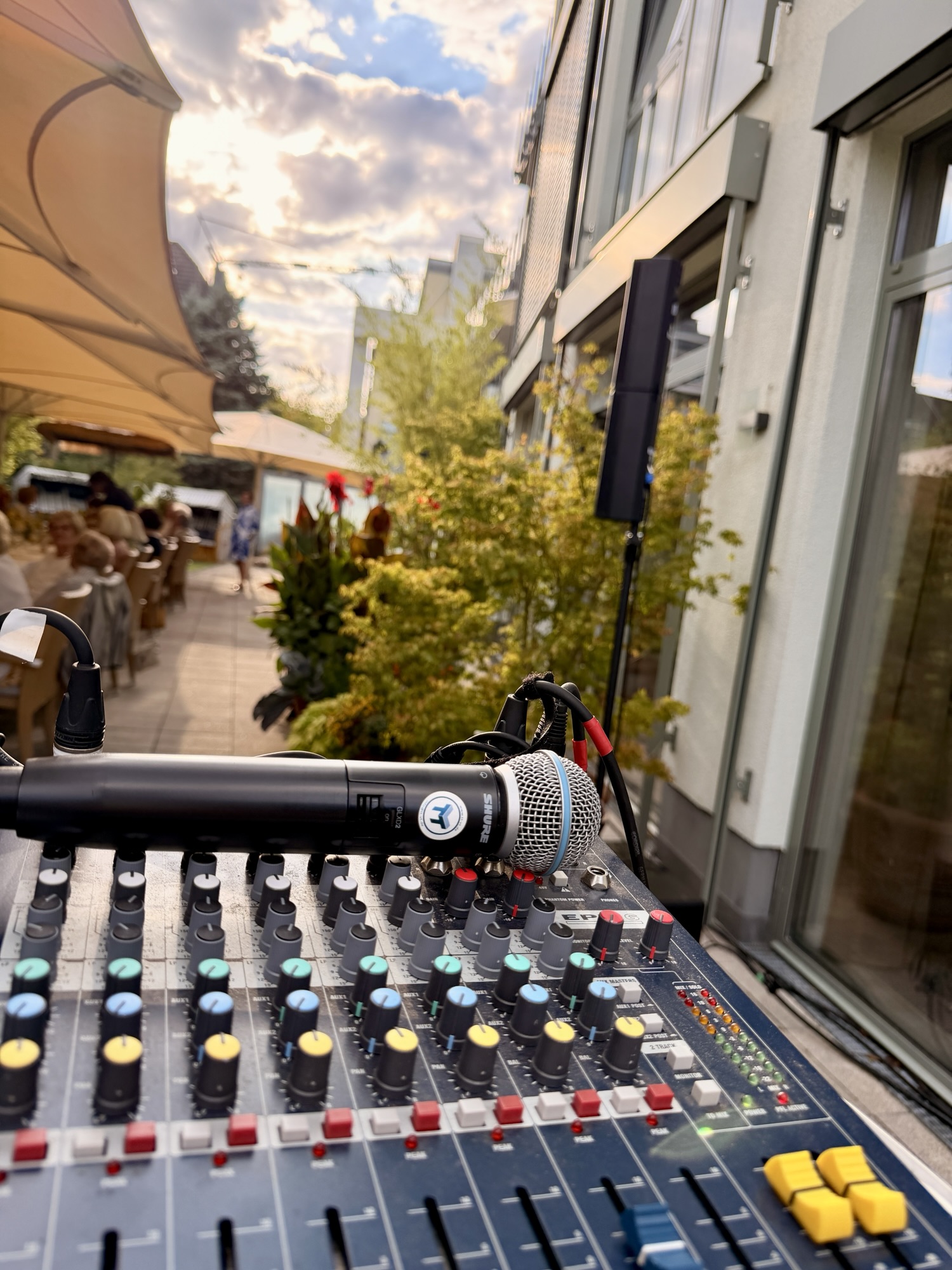 Blick über ein Mischpult mit Shure-Handmikrofon auf eine Restaurant-Terrasse mit Sonnensegeln bei Abendlicht, im Hintergrund die Fassade des Gebäudes